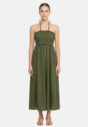 Donna che indossa un vestito midi verde oliva con collo halter, corpetto arricciato e sandali bassi beige con cinturini, in piedi su sfondo bianco.