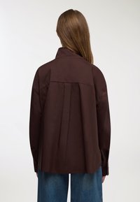 Camicia marrone con bottoni, pince sul retro, maniche lunghe e colletto, indossata sopra jeans larghi color azzurro chiaro. Tessuto dalla texture liscia.