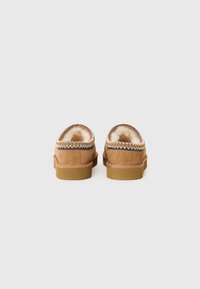 Pantofole in suede marrone con fodera in shearling, punta arrotondata e cuciture decorative lungo il bordo. Suole in gomma piatta per una buona aderenza.