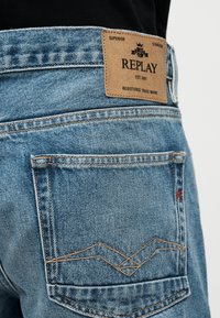 Κοντινό πλάνο στην πίσω τσέπη και το καφέ δερμάτινο έμβλημα της μάρκας Replay σε ανοιχτό μπλε τζιν τζιν, φορεμένα με μαύρο τοπ.