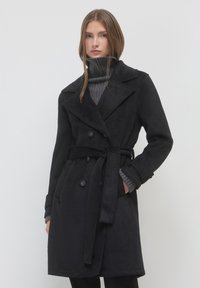 Donna che indossa un cappotto nero doppiopetto con cintura sopra un maglione grigio a collo alto, in piedi davanti a uno sfondo bianco semplice.