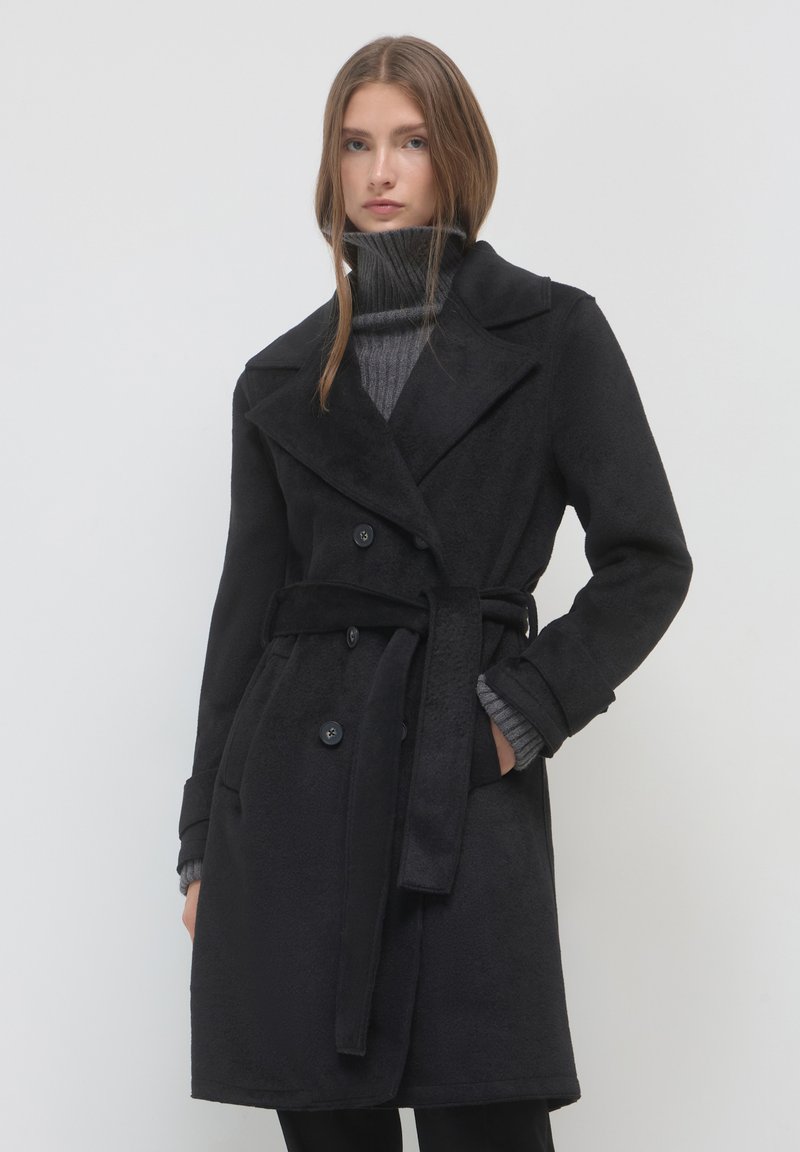 Donna che indossa un cappotto nero doppiopetto con cintura sopra un maglione grigio a collo alto, in piedi davanti a uno sfondo bianco semplice.