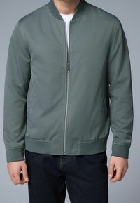 Chaqueta bomber en tejido verde oscuro con cremallera frontal, cuello y puños acanalados, que presenta un diseño minimalista y dos bolsillos laterales.
