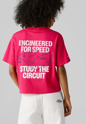 Personne aux cheveux bouclés portant un t-shirt court rose montrant une carte de circuit de course avec le texte "Conçu pour la vitesse, étudiez le circuit."