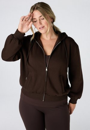 Femme aux cheveux blonds portant un sweat à capuche marron foncé zippé et un pantalon assorti, levant la main droite près de son visage.