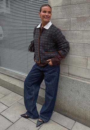 Bruine gestreepte cropped jas met een witte kraag, gecombineerd met wijde donkere spijkerbroeken en puntige zwarte schoenen, staand tegen een gestructureerde muur.