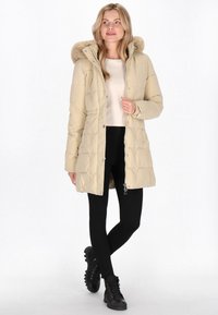 Beige pufferjakke med en pelsforet hætte, lynlås og trykknaplukning, quiltet tekstur, båret med sorte leggings og sorte snørebåndsstøvler.