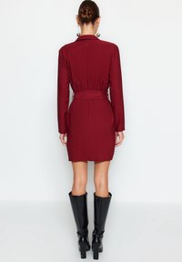 En bordeauxklänning med bälte i midjan, långa ärmar och krage. Knälång design med en slät yta, matchad med knähöga svarta stövlar.