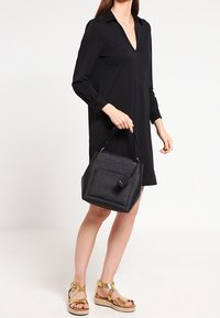 Czarna torebka o uporządkowanej formie, w kształcie prostokąta, z gładką powierzchnią i odczepianą metką. Złote akcenty wykończenia.