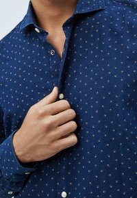 Laivastonsininen nappipaita, jossa on pieniä toistuvia valkoisia kuvioita. Siinä on terävä kaulus, yksittäinen rintatasku ja helmiäisnapit.