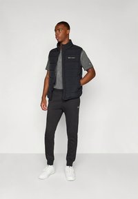 Crna podstavljena prsluk preko sive majice kratkih rukava, uparena s crnim trenirkom i bijelim tenisicama. Jednostavan dizajn s vidljivim logotipima.