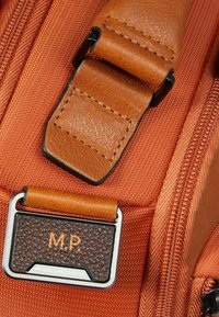 Cinturino in pelle marrone con un dettaglio testurizzato, fissato a una base in tessuto arancione con un'insegna incisa e un dettaglio in metallo argentato.