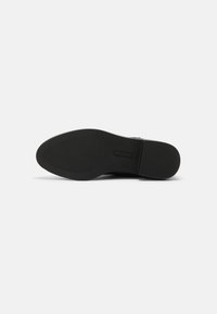 Suela de zapato negra con patrón de textura, tacón plano y nombre de la marca grabado cerca del área del arco, mostrado sobre fondo blanco.