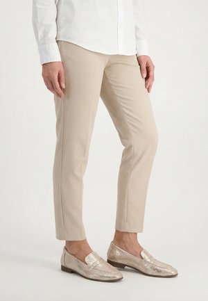 Kvinde iført beige slim-fit bukser og metallicmønstrede loafers, med en hvid langærmet skjorte med knapper synlig ved taljen.