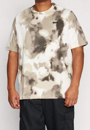 Homme portant un t-shirt tie-dye beige et noir à manches courtes avec un petit logo Calvin Klein sur la poitrine, assorti à un pantalon noir.