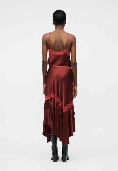 Vrouw die een mouwloze midi-jurk van rode satijn draagt met kanten afwerking en een asymmetrische zoom, gecombineerd met zwarte laarzen met hak, staand met de rug naar de camera.