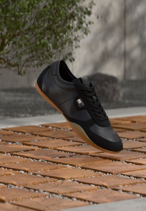 Čierna kožená teniska s podrážkou z gumy vznášajúca sa nad drevenou dlažbou vonku pri šedej stene a zelenej vegetácii.