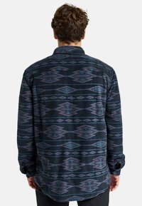 Camicia di flanella con una base blu scuro, caratterizzata da un motivo geometrico in viola e teal. Maniche lunghe, colletto e design con orlo curvo.