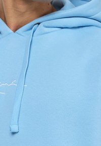 Sudadera azul claro de algodón suave, con un bolsillo tipo canguro en la parte frontal, cordones y un sutil detalle bordado en el pecho.