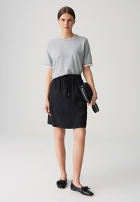 Junge Frau in einem grauen kurzärmligen Shirt, das in einen schwarzen knielangen Rock gesteckt ist, hält eine schwarze Clutch und steht vor einem schlichten Hintergrund.