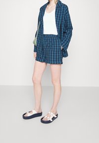 Blå och svart rutiga kortärmade skjorta med matchande shorts; vit linne; platåsandaler med en ljus ovandel och mörk sula; grön axelväska.