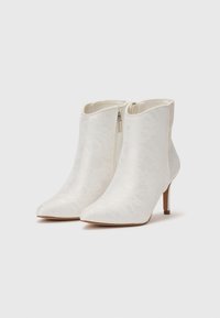 Paire de bottines blanches avec fermetures éclair latérales, bouts pointus et talons hauts fins sur un fond clair uni.