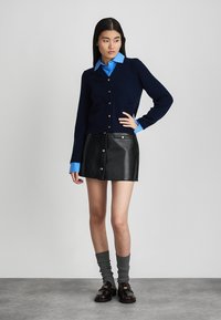 Ναυτικό πλεκτό cardigan με μπροστινές τσέπες, ανοιχτό μπλε πουκάμισο από κάτω, μαύρη δερμάτινη μίνι φούστα, γκρι κάλτσες μέχρι το γόνατο και σκούρα loafers.