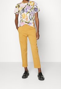 Camiseta de algodón con estampado floral en tonos púrpura, amarillo y naranja, combinada con pantalones ceñidos en color mostaza y mocasines negros de suela gruesa.