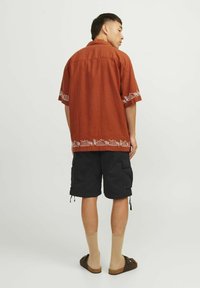 Korte mouw, roestkleurig shirt met een wit geborduurd patroon aan de zoom, gecombineerd met zwarte cargoshorts en beige kniekousen.
