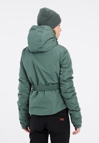 Gevoerde jas in een gedempt groen, met een capuchon en een elastische tailleband. Draag met een bijpassende gebreide muts, weergegeven vanuit de achterkant.