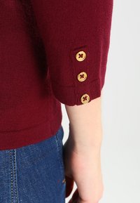 Maglione color bordeaux con tre bottoni dorati al polsino. Tessuto in maglia morbida, con dettagli a coste all'orlo e alle maniche.