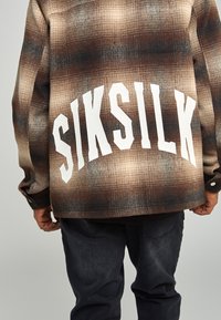 Brun och beige rutig flanellskjorta med vitt "SIKSILK"-logotyp på ryggen. Har en avslappnad passform och knäppning på  ärmarna.