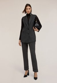 Chaleco gris texturizado a medida y pantalones a juego con dobladillo. Top negro de manga larga debajo y bolso negro. Tacones altos negros.