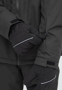 Mustad kindad kohandavad musta jope varrukat. Jope on sileda tekstuuriga, varustatud tõmblukuga ja Velcro rihmaga varruka sulgemiseks.