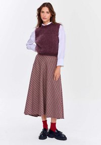 Fialová bezrukávová svetr přes bílou košili, doplněná dlouhou plisovanou sukní s červeným a černým kostkovaným vzorem. Černé boty s tlustou podrážkou.