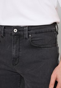 Czarne dżinsy o dopasowanym kroju ze stretchem, z klasycznym pięcioma kieszeniami, zapięciem na guziki i widocznym przeszyciem. Noszone z białą koszulą.