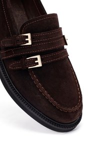 Mocassino in suede marrone con dettagli a doppia fibbia, cuciture a contrasto, punta rotonda e suola in gomma. La vista ravvicinata mette in risalto la texture e il design.