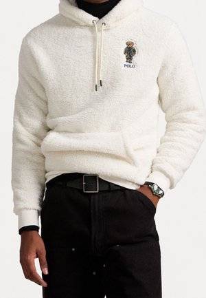 Weißer, flauschiger Hoodie mit Kängurutasche, Kordelzug und besticktem Bären mit schwarzer Jacke über dem Schriftzug "POLO", kombiniert mit schwarzer Hose und Uhr.