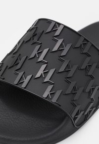 Sandale noire à glissière avec une sangle texturée ornée d'un motif en relief du logo "kl" et un coussin de pied lisse et mat. Design léger et flexible.