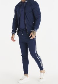 Mężczyzna ubrany w granulowany granatowy dres z białymi paskami, do tego białe trampki, stoi na białym tle.