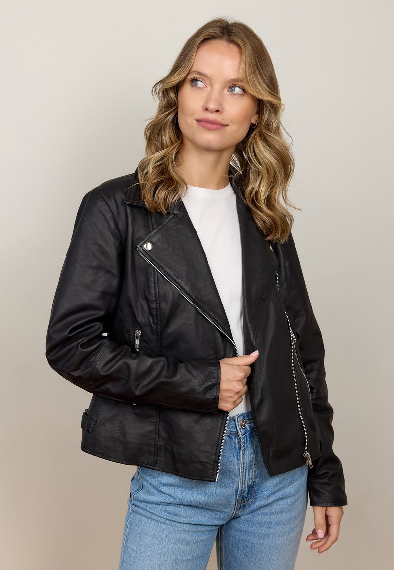 Schwarze Leder-Motorradjacke mit silbernen Beschlägen, Reißverschlusstaschen und einer leicht verkürzten Passform, getragen über einem weißen Hemd und hellblauen Jeans.