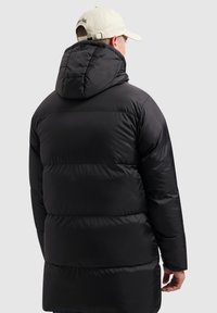 Man draagt een lang zwart pufferjack met een capuchon en een beige baseballpet, van achteren gezien tegen een effen lichte achtergrond.