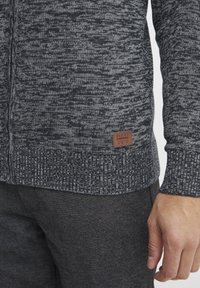 Maglione lavorato a maglia grigio scuro con motivo testurizzato, polsini e orlo a coste. Include una piccola etichetta in pelle marrone sul fondo.