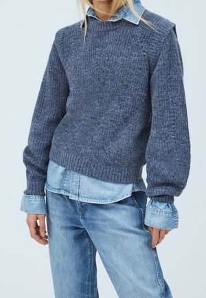 Person iført lagdelt blå striktrøje over lyseblå denimskjorte med blå jeans mod en ensfarvet lys baggrund.