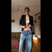Blazer nero sopra un top bandeau bianco, abbinato a jeans di denim azzurri a gamba larga. La stanza presenta un divano e luci sospese.