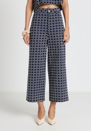 Pantaloni larghi cropped blu navy con motivo a catena geometrica bianca, vita alta, bottone frontale, indossati con top abbinato e tacchi beige a punta.