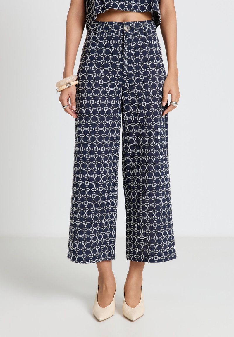Navy wide-leg cropped pants with white geometric chain pattern, high waist, front button, worn with matching crop top and beige pointed heels.