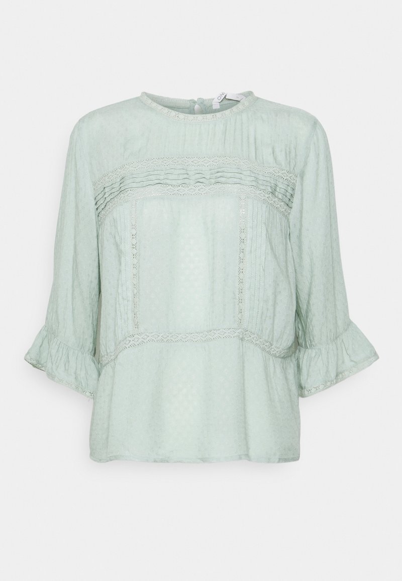 Blouse vert clair à manches 3/4, bordures en dentelle, plis verticaux et poignets volantés, avec encolure ronde et fermeture par boutons dans le dos.