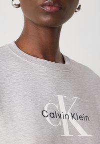 Gray cotton sweatshirt featuring "Calvin Klein" logo in white and black embroidery. Close-up view of neckline and textured fabric.