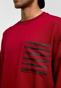 Rode sweatshirt met een borstzak en zwart gestreepte details. Het materiaal is zacht en de textuur lijkt glad met subtiele stiksels.